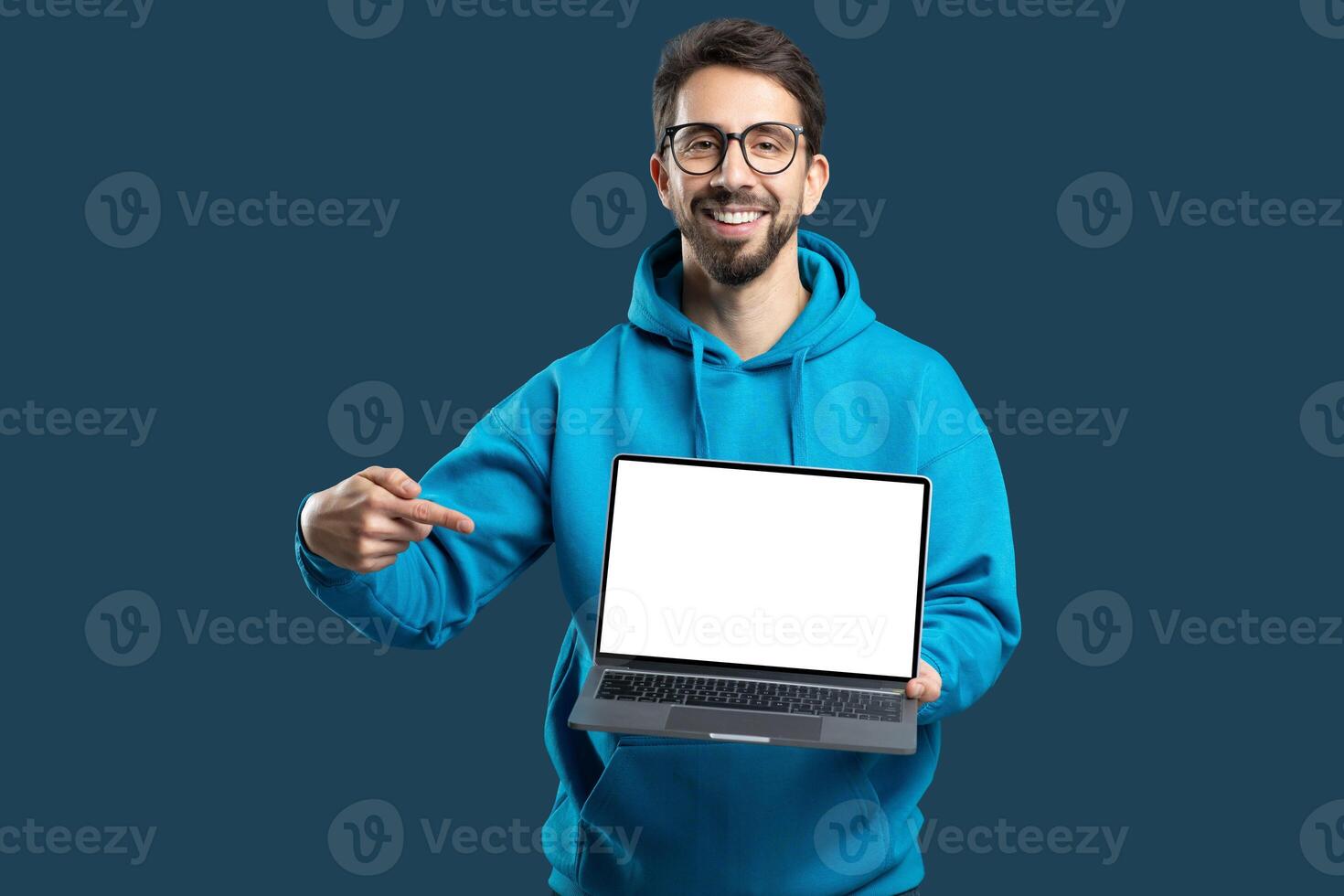 A cheerful young man wearing a bright blue hoodie stands against a solid background while presenting a blank laptop screen. He points towards the laptop, inviting attention and engagement photo