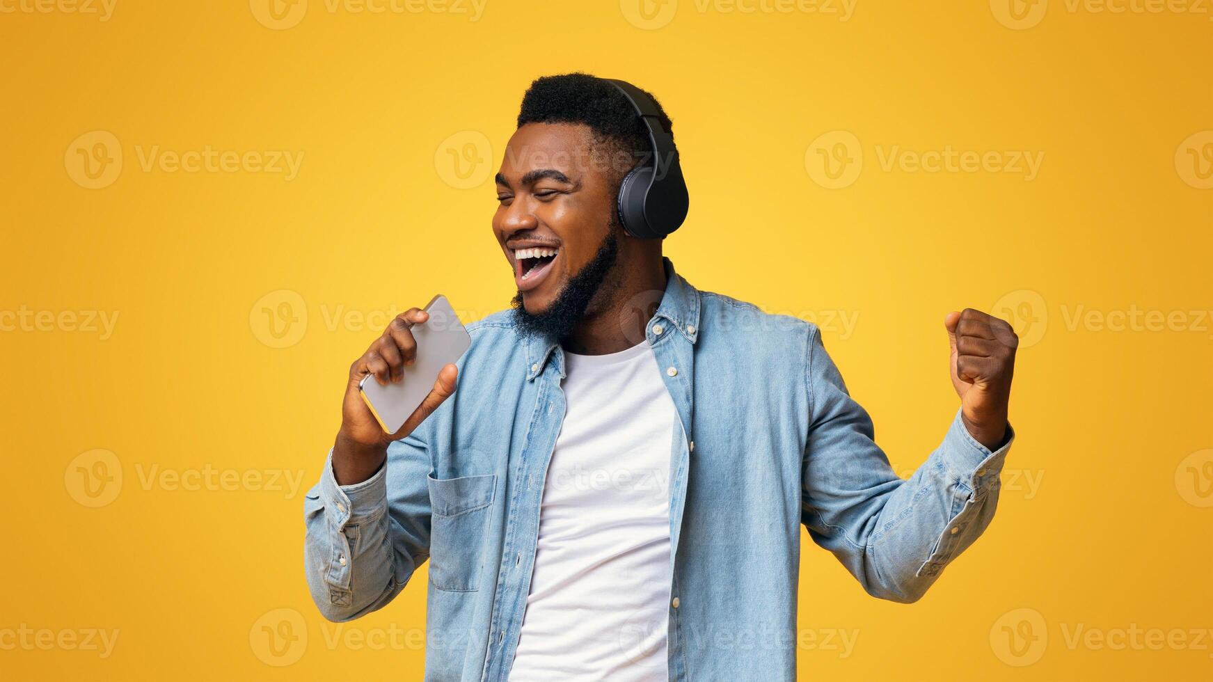 Joyful african american man in wireless headphones singing into smartphone like microphone on yellow background, panorama photo
