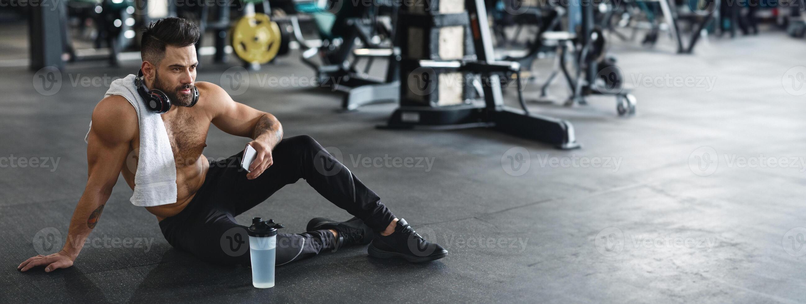 un joven hombre se sienta en el gimnasio piso, tomando un descanso después hacer ejercicio, con un toalla alrededor su cuello y un agua botella cercano, Copiar espacio foto