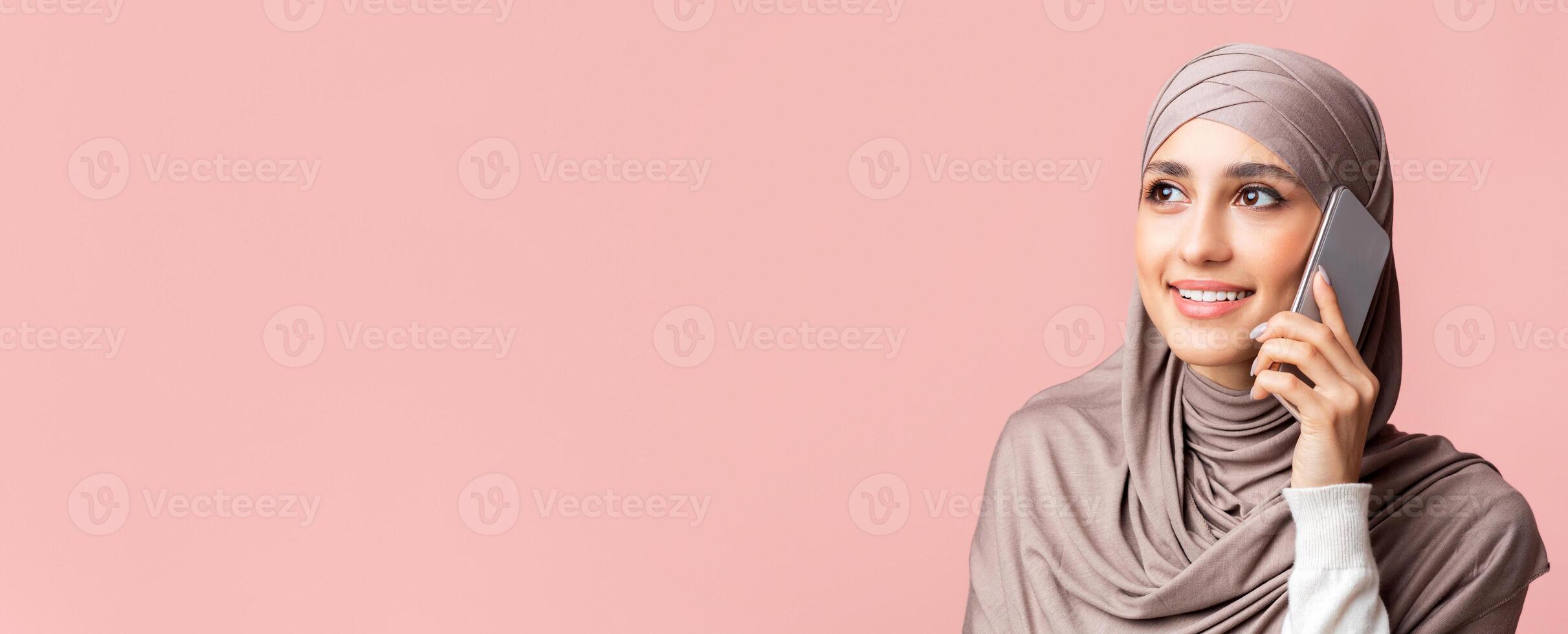 Always in touch. Beautiful muslim girl in hijab talking on cellphone and looking away over pink background with empty space, closeup photo