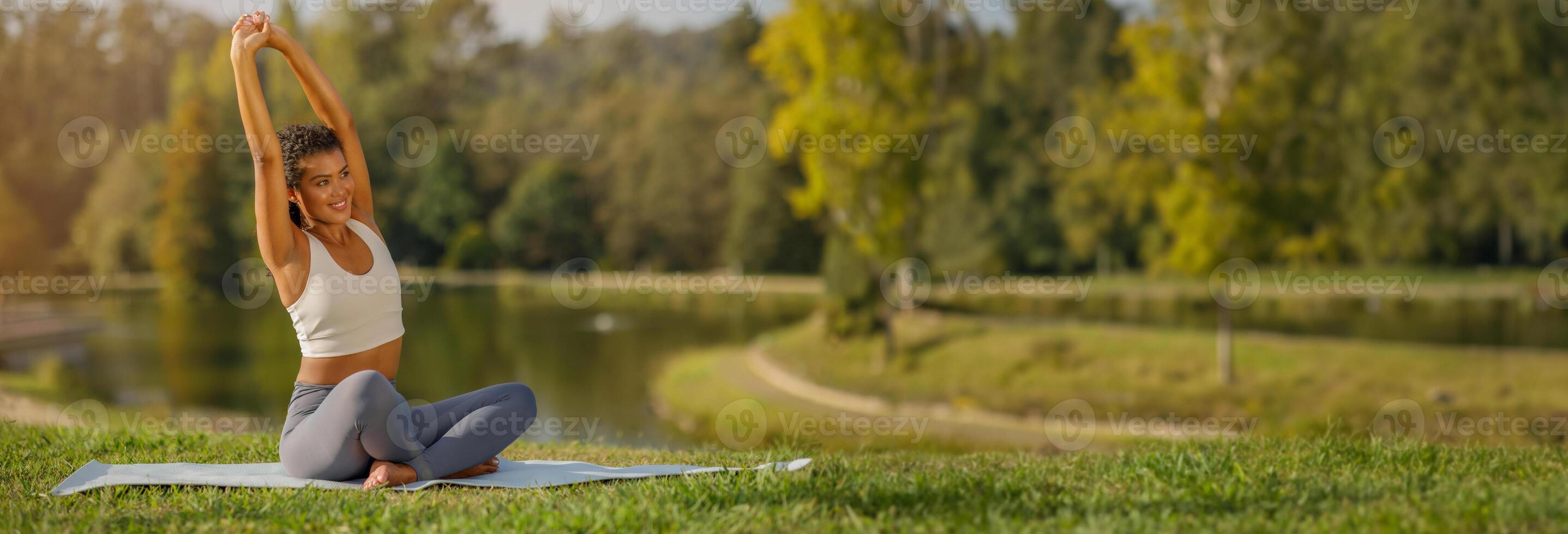 Sportswoman practicing pilates outdoors, bending in half lotus pose with arms extended, epitomizing health and flexibility, sitting on mat in green park, copy space photo