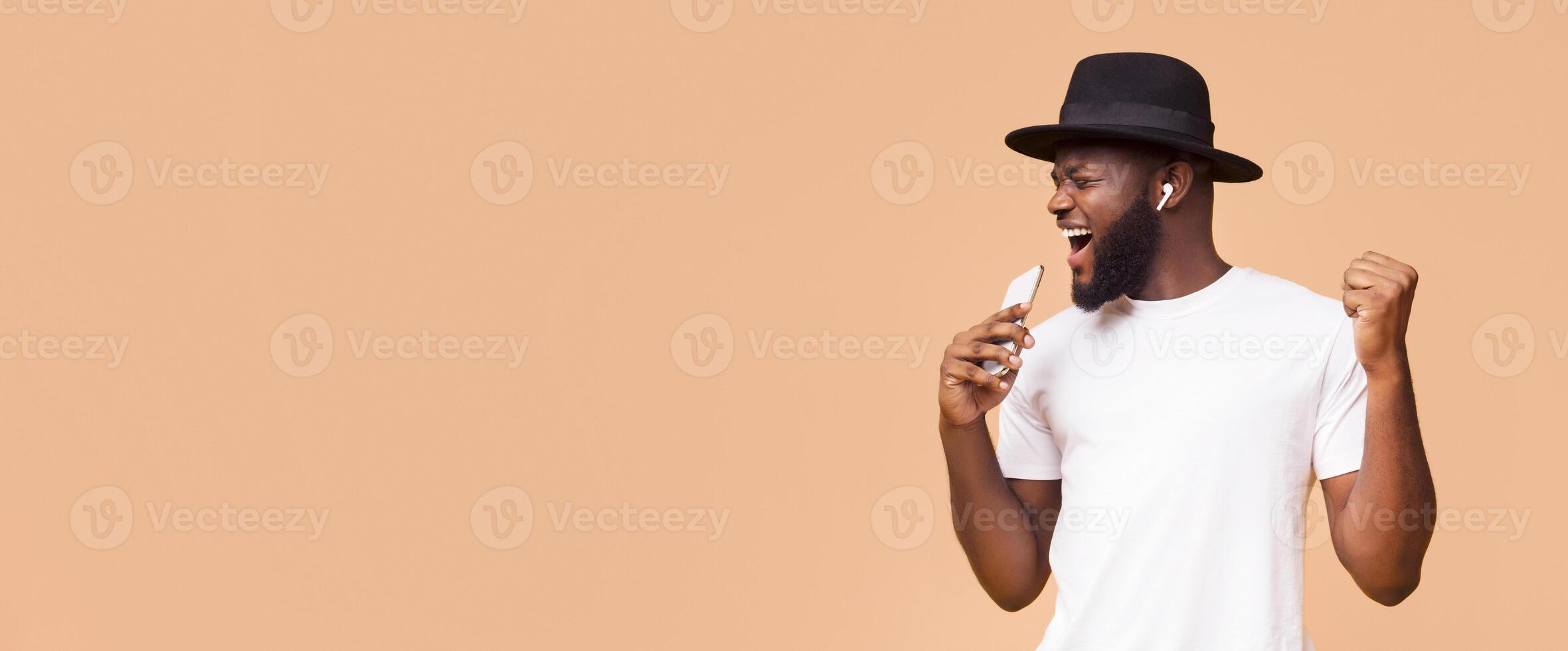 African American man with a beard and a black hat is singing into his phone. His right hand is raised in the air, and he is smiling and looking at copy space photo