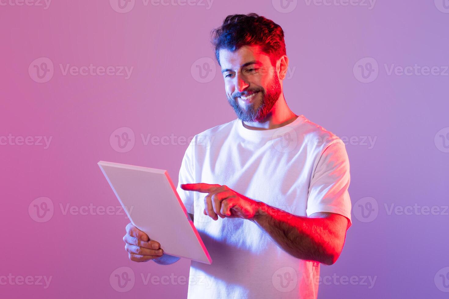 A bearded man wearing a white t-shirt interacts with a digital tablet while standing in a studio with purple and pink neon lighting. He is smiling and appears engaged with the content on the tablet. photo