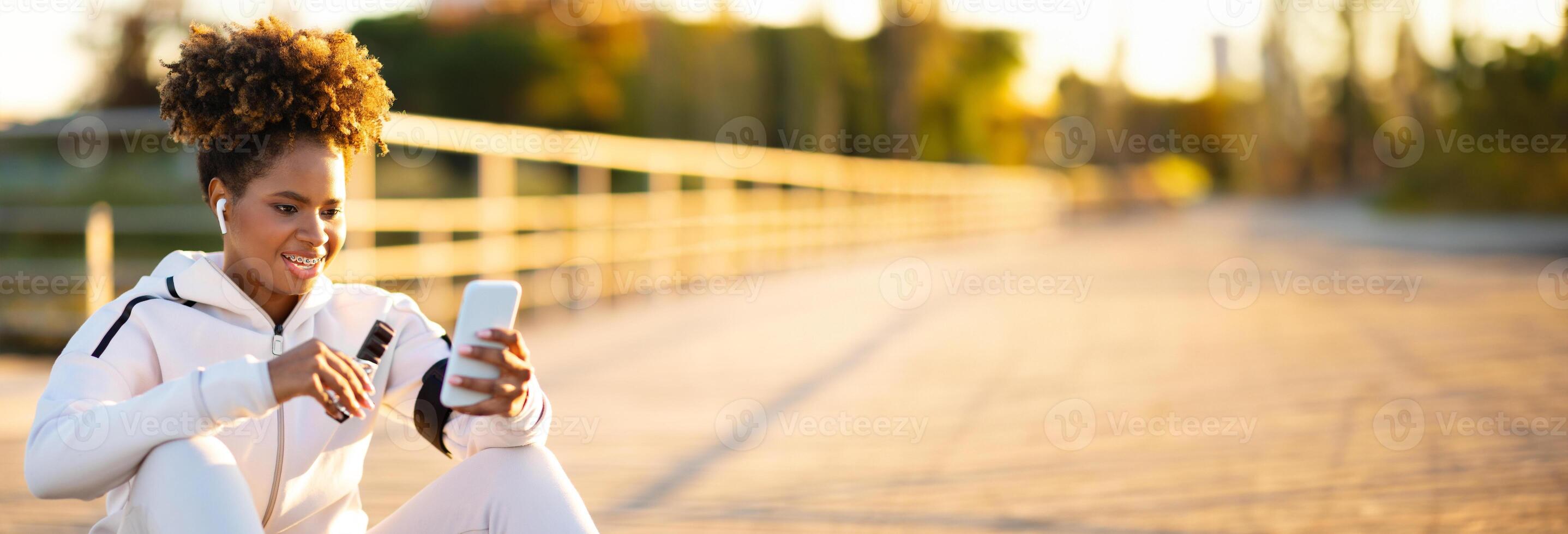 Snack Break. Young Black Woman Resting Outdoors After Sport Training, Smiling African American Lady Using Smartphone And Eating Protein Bar While Relaxing After Jogging Outside, Copy Space photo