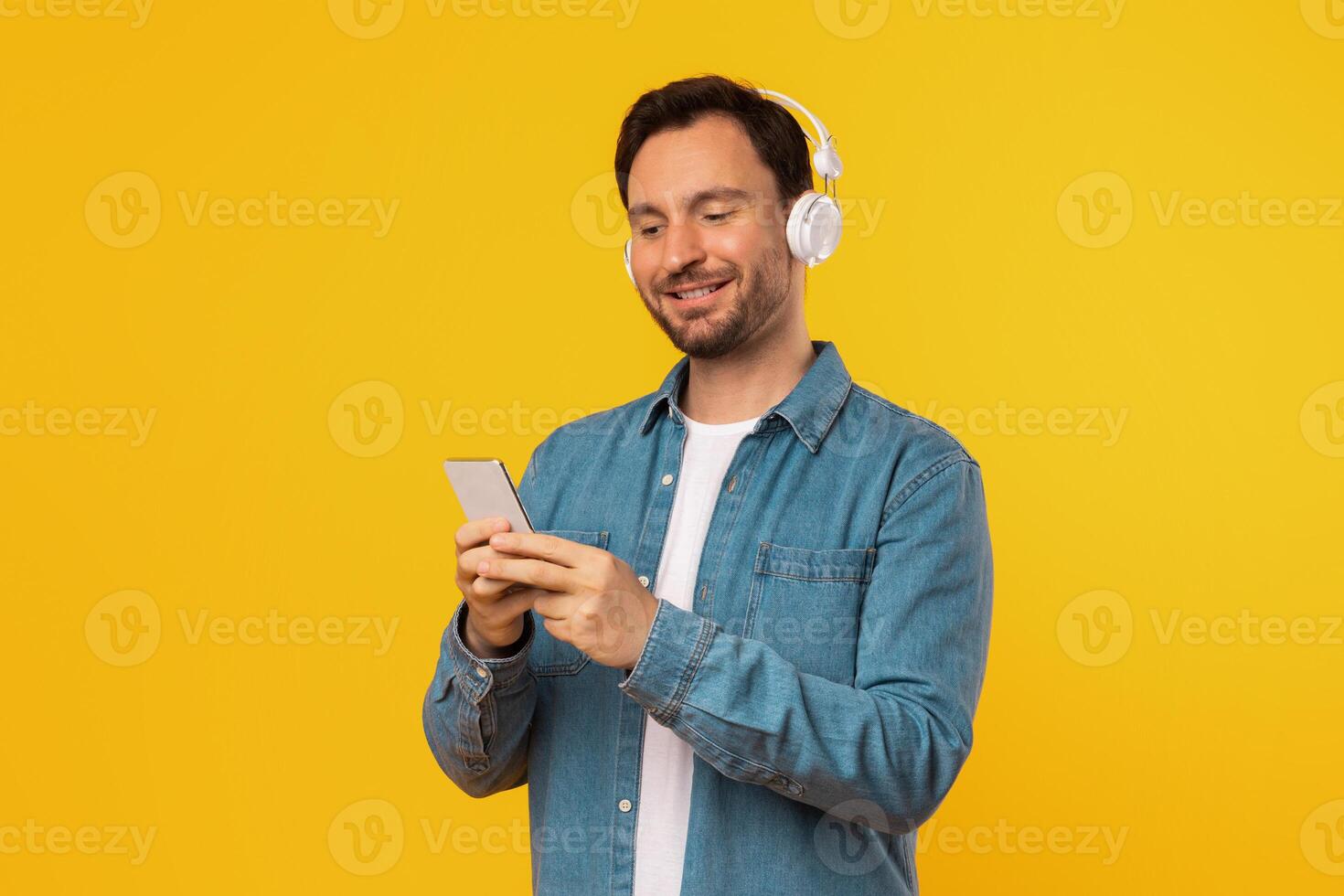 A man with a beard is standing in front of a yellow background. He is wearing a blue denim shirt and headphones. He is smiling and looking at his smartphone. photo