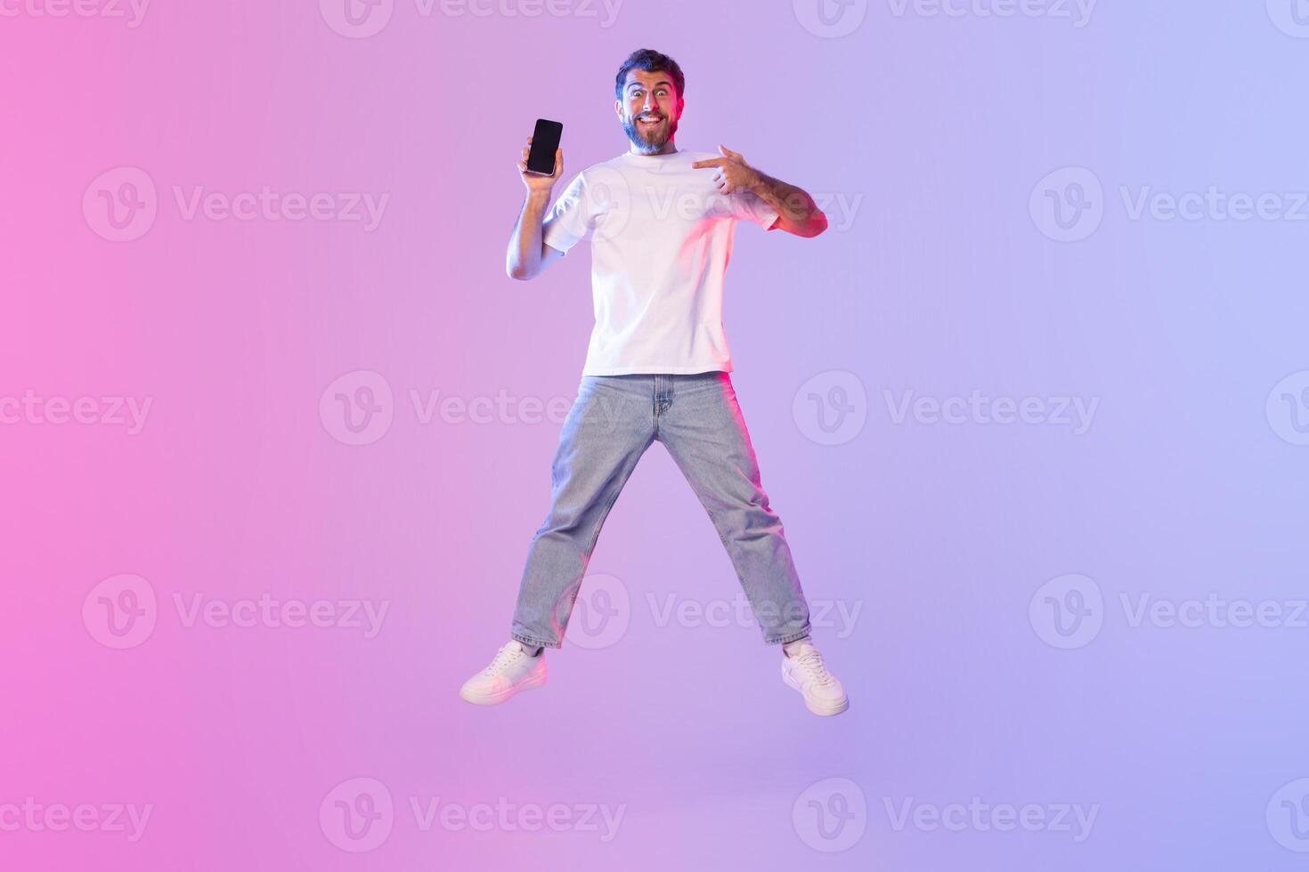 A young man dressed in a white t-shirt and jeans energetically jumps while holding and pointing at his smartphone. The backdrop features an eye-catching gradient of pink and blue photo