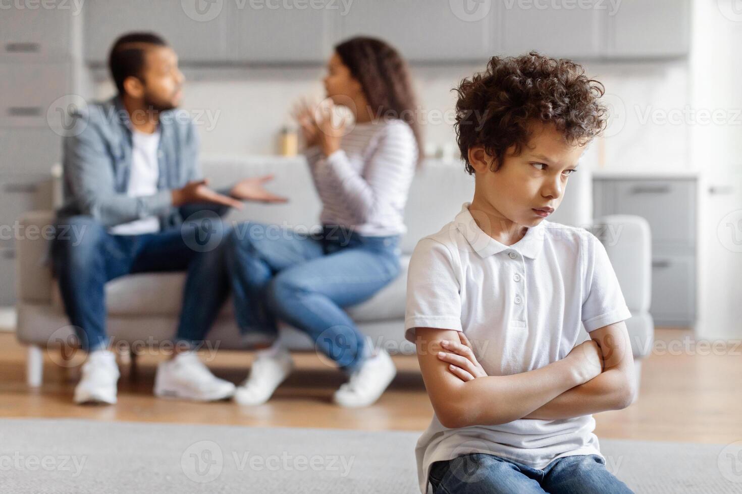 African American young boy stands in the foreground with arms crossed and a frown, displaying feelings of unhappiness or frustration, parents appear to be having a heated conversation photo