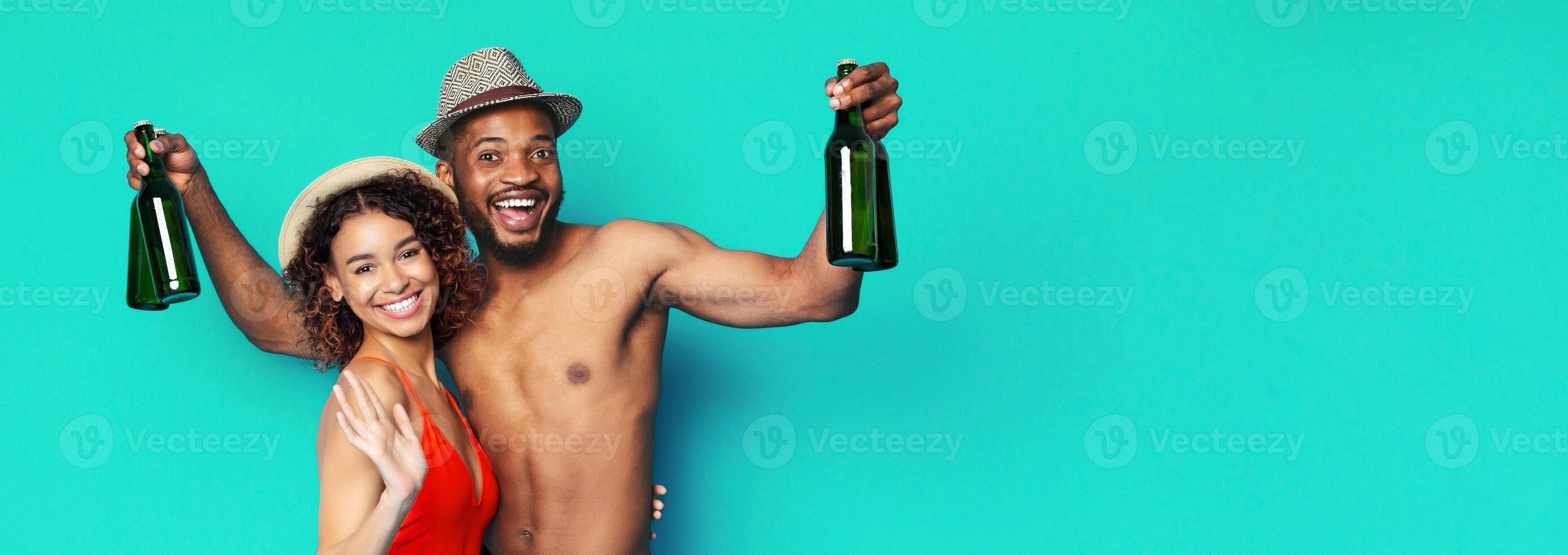 African American couple is smiling and holding bottles in front of a bright turquoise background. The man is shirtless and wearing a hat, and the woman is wearing a red swimsuit and a straw hat. photo