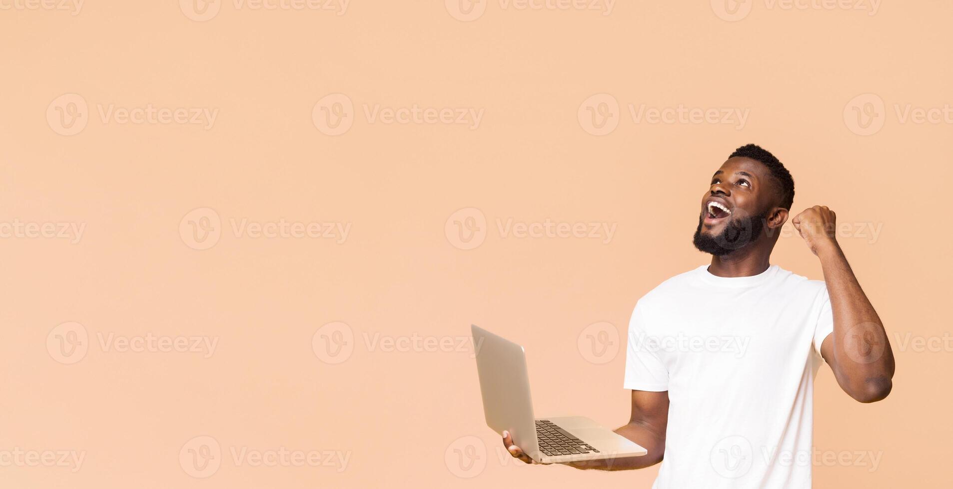An African American man in a white t-shirt is holding a laptop computer with his left hand and raising his right fist in the air, looking upwards with a joyous expression, copy space photo
