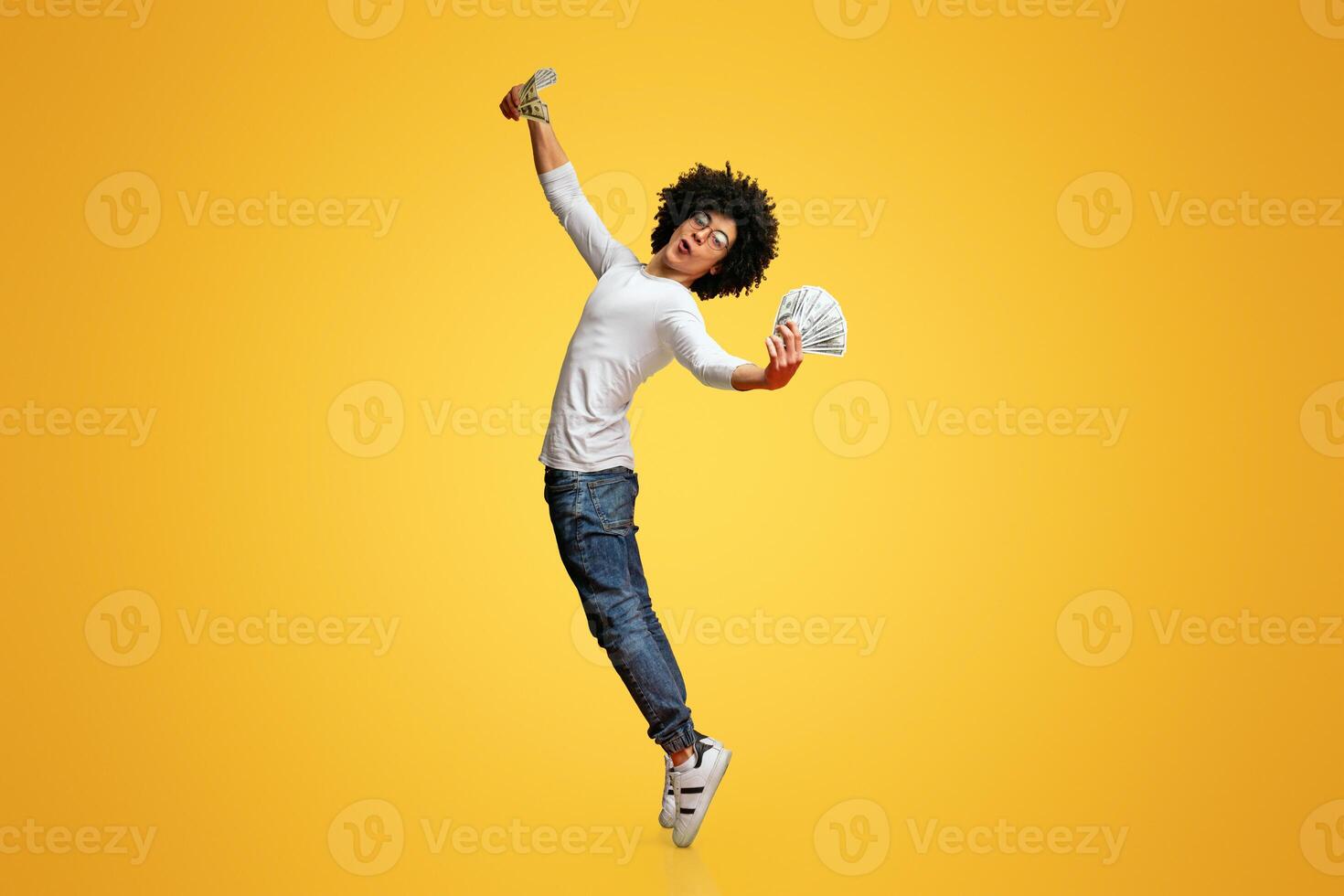 Young excited black bushy guy dancing with dollars, standing on tiptoes with money fans, orange studio background photo