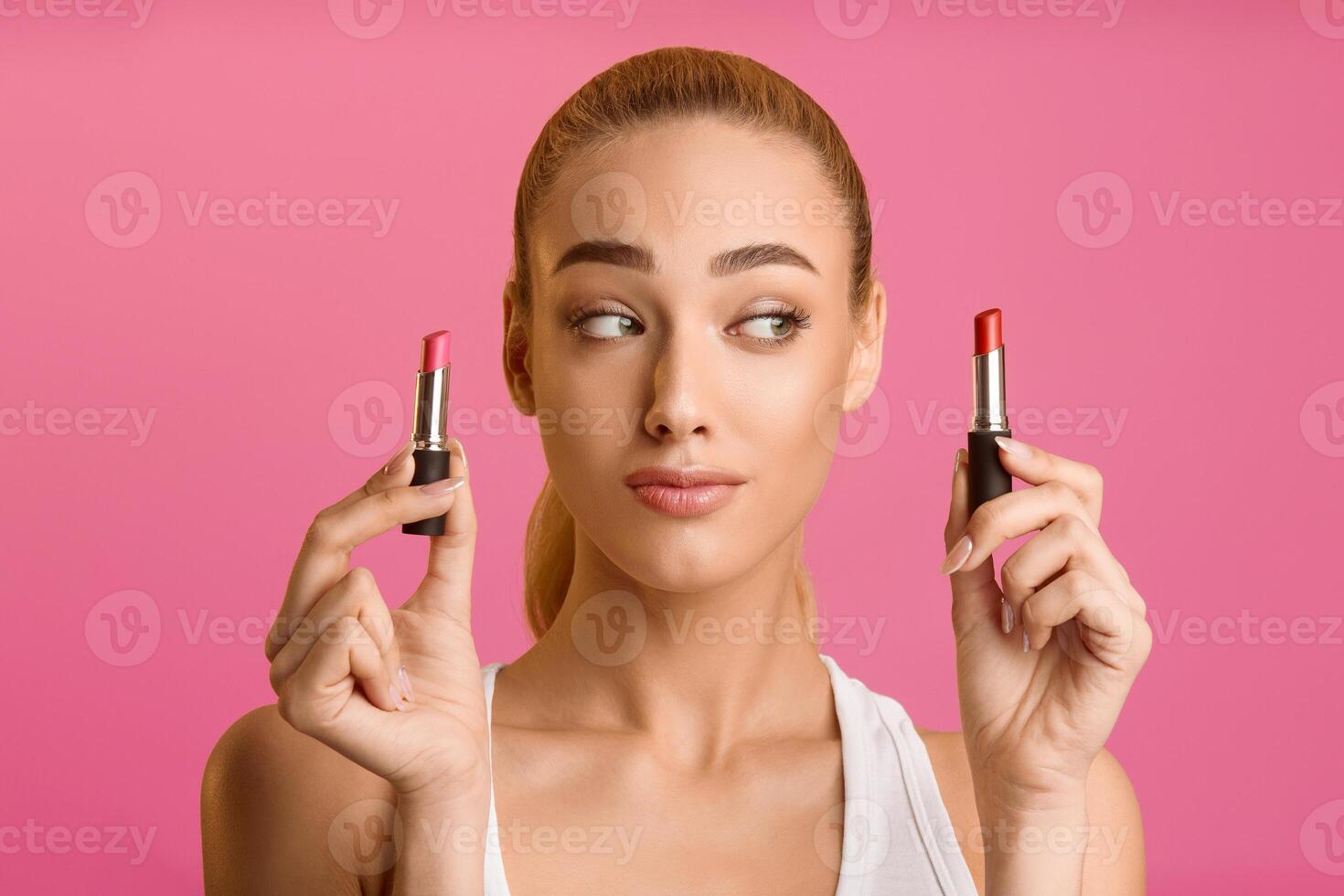 A young woman with blonde hair stands in front of a pink background. She is holding two lipsticks, one in each hand, and she is looking to the side with a thoughtful expression. photo