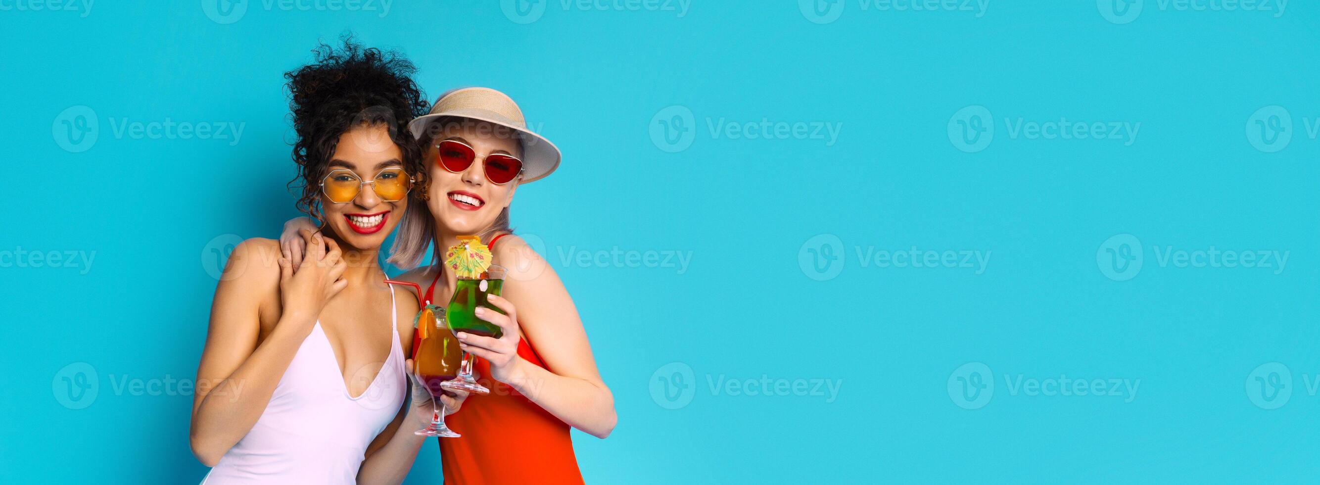 Two young women smiling at the camera. The woman on the left is wearing a white bathing suit and yellow sunglasses, while the woman on the right is wearing a red dress and a hat, copy space photo