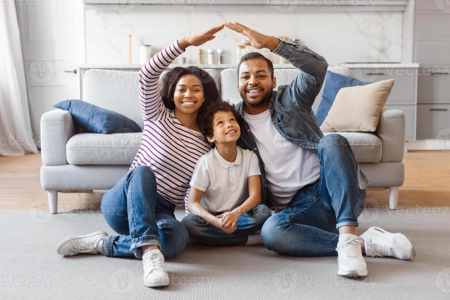 A cheerful African American family consisting of a mother, father, and child are sitting on the floor in a well-lit living room. They are forming roof with their arms above them photo