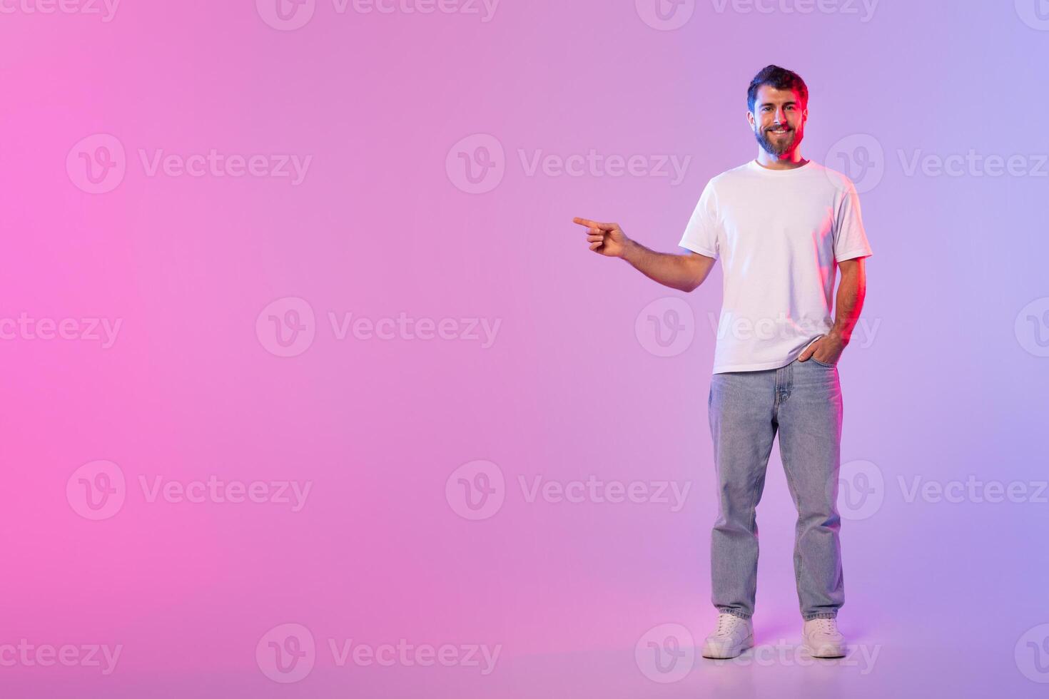 A man wearing a white t-shirt and jeans stands casually with one hand in his pocket, pointing with the other hand at copy space. The background features a gradient of pink and purple studio lighting photo