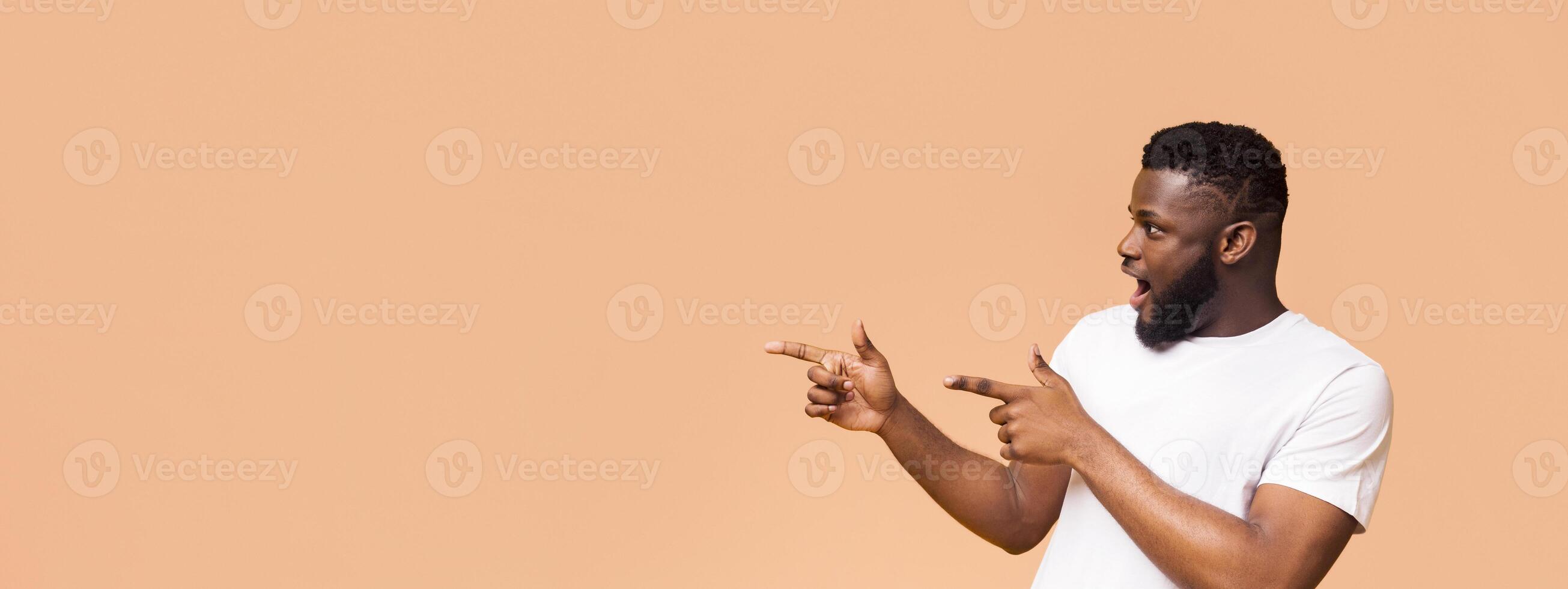 This image shows African American man with a beard and a white shirt, looking surprised and pointing to the right. He is standing in front of a peach-colored background, copy space photo