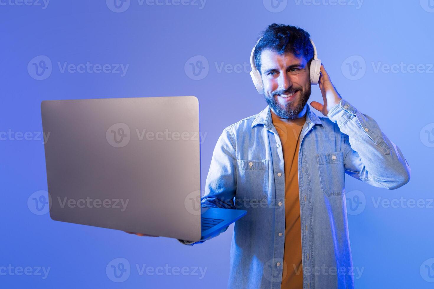 A cheerful young man is holding a laptop in one hand while listening to music with headphones. Background is illuminated with soft blue lighting, indicating he is likely enjoying a nighttime session photo