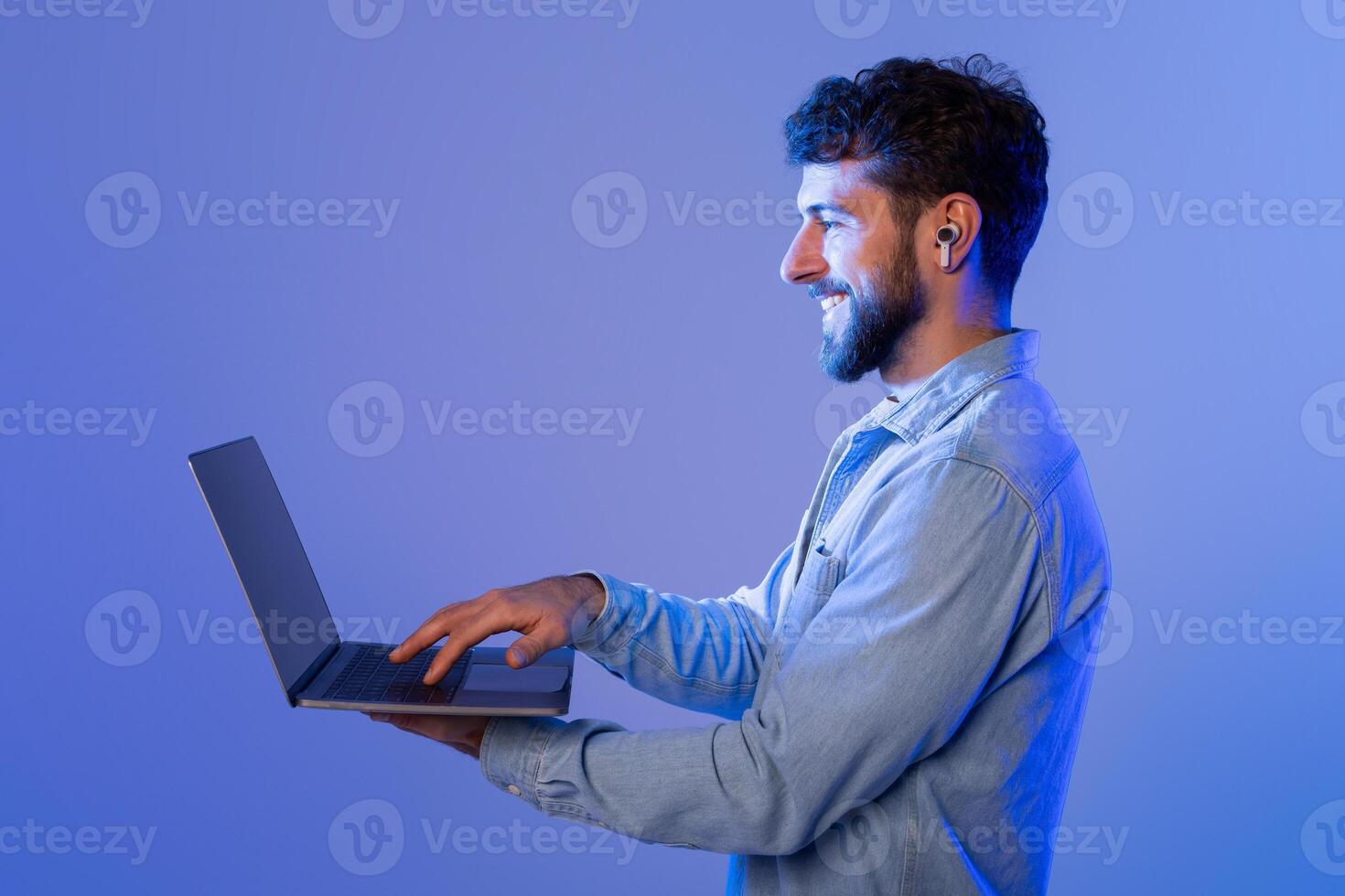 A man with a beard is sitting with a laptop open in front of him. He appears focused as he types on the keyboard, possibly working or studying. photo