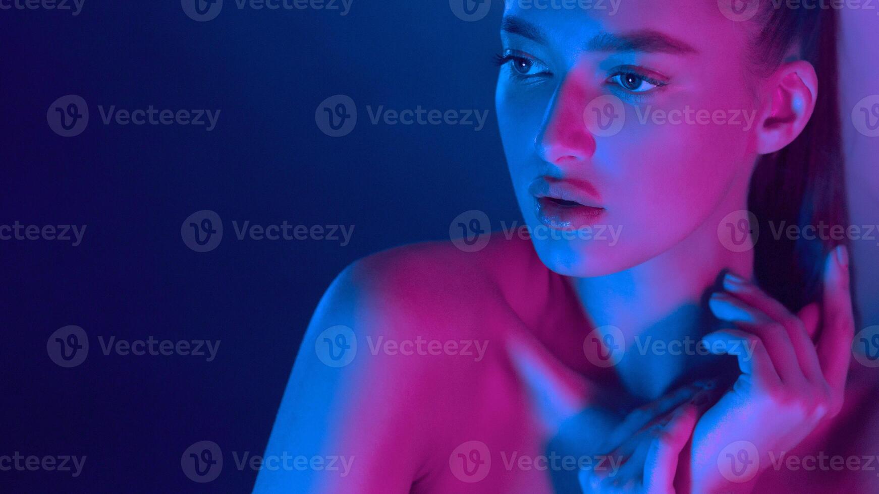 A woman gazes thoughtfully into the distance, bathed in vibrant neon lights that blend blue and pink hues, creating a striking and contemplative scene. photo