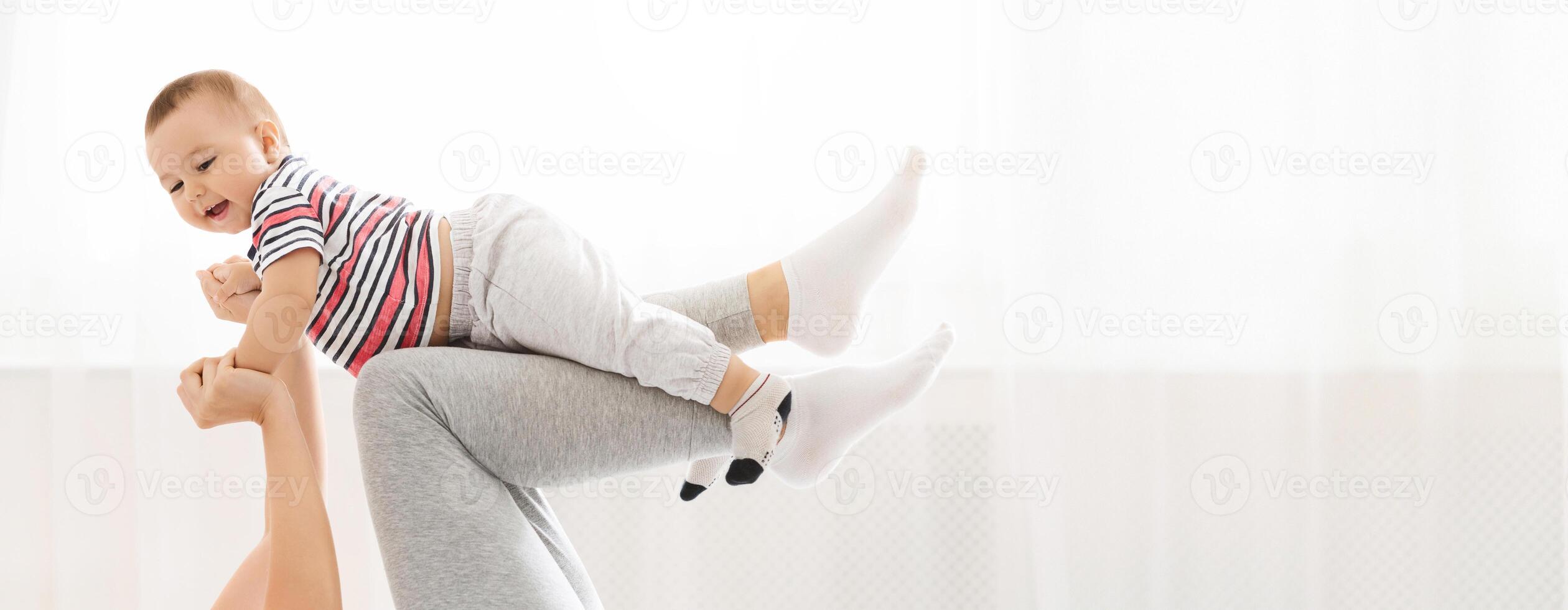 A caregiver joyfully lifts a smiling baby into the air during a playful moment indoors. The child appears delighted, while the caregiver expresses warmth and affection, creating a vibrant atmosphere photo