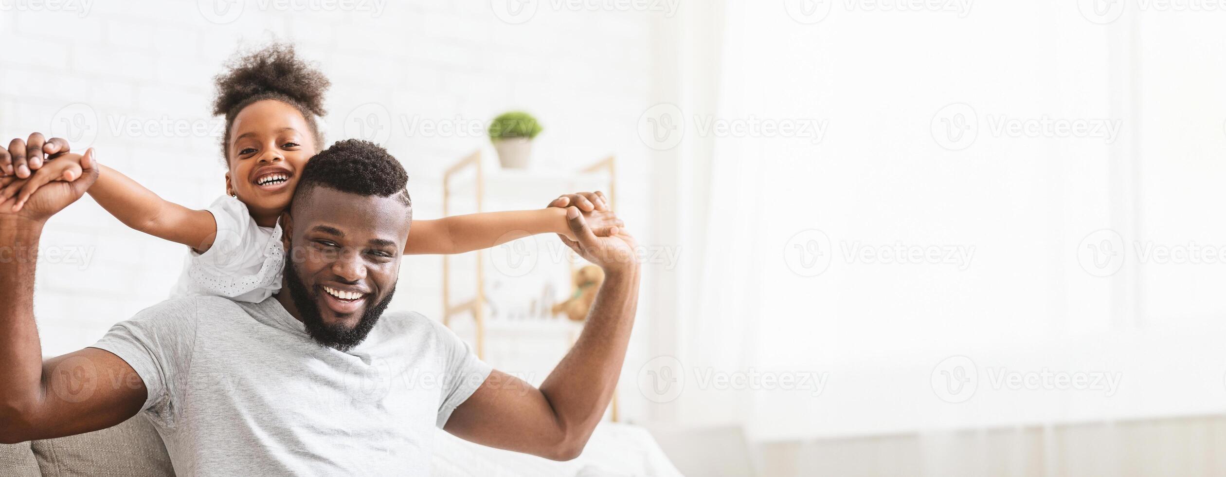 A delighted black father shares a playful moment with his young daughter in a well-lit living room. The little girl sits on her father shoulders, copy space photo