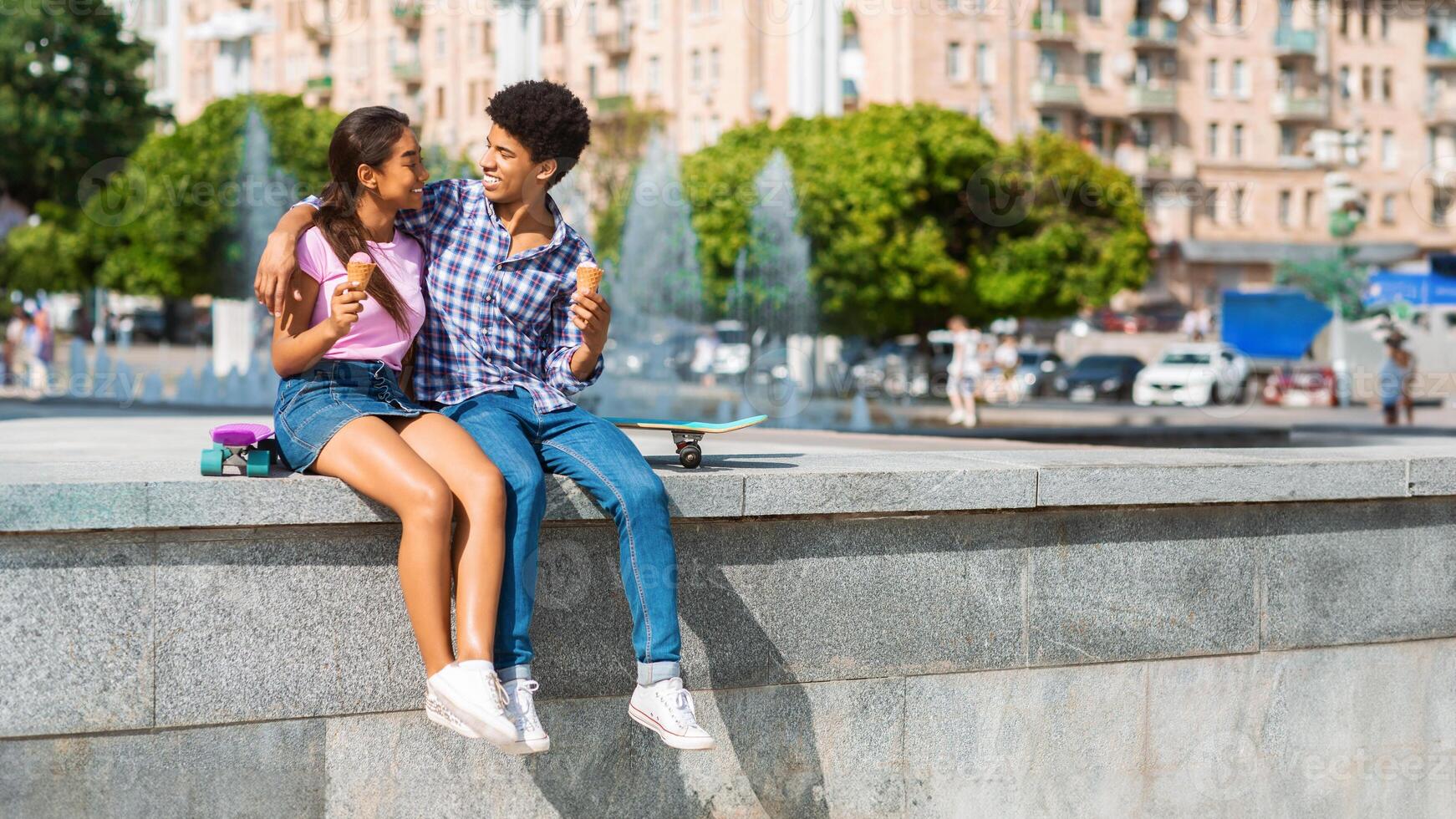 This image shows a teen black couple sitting on a wall next to fountain in a city park, enjoying ice cream cones and looking at each other, copy space photo