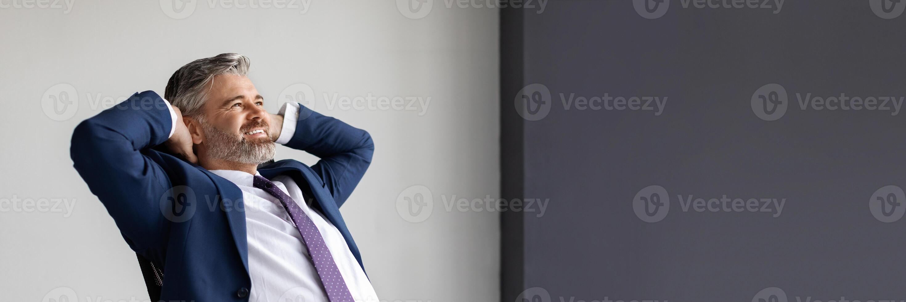 Smiling middle aged businessman leaning back in chair at workplace in office, handsome happy entrepreneur in suit sitting at desk with hands behind head, resting after successful day, copy space photo