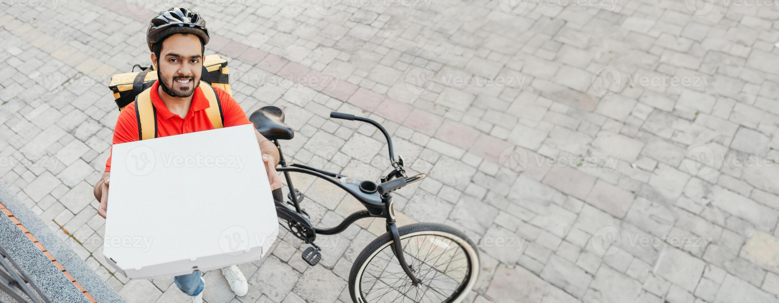 Order delivery. Smiling young man with beard, in helmet, shows box of pizza to peephole, near bike on street, free space photo