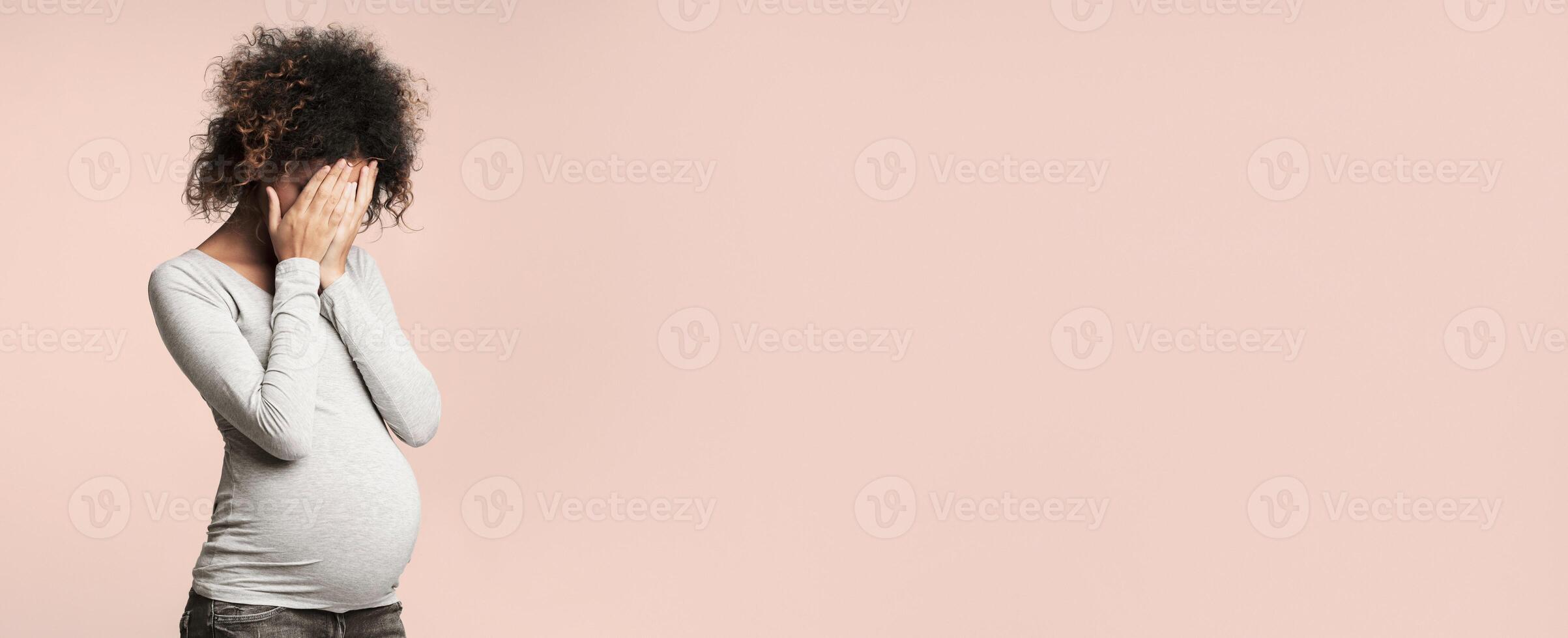 Depression during pregnancy. Afro woman covering face with hands and crying, grey background photo