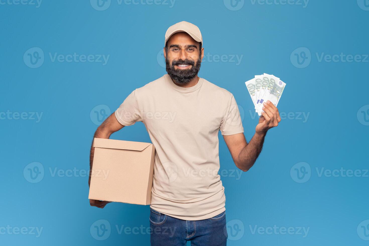 Indian delivery man stands against a blue background in a studio, smiling and holding a cardboard package in one hand and paper banknotes in the other photo