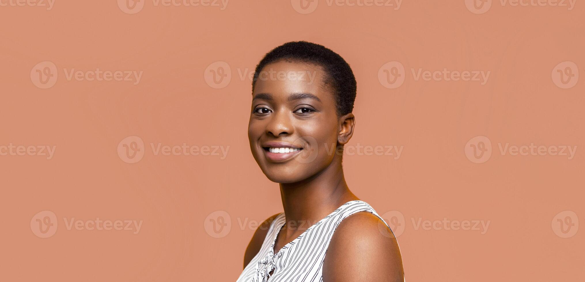A portrait of a smiling Black woman with short hair. She is wearing a white and black striped tank top and is looking at the camera. The background is a peach color. photo
