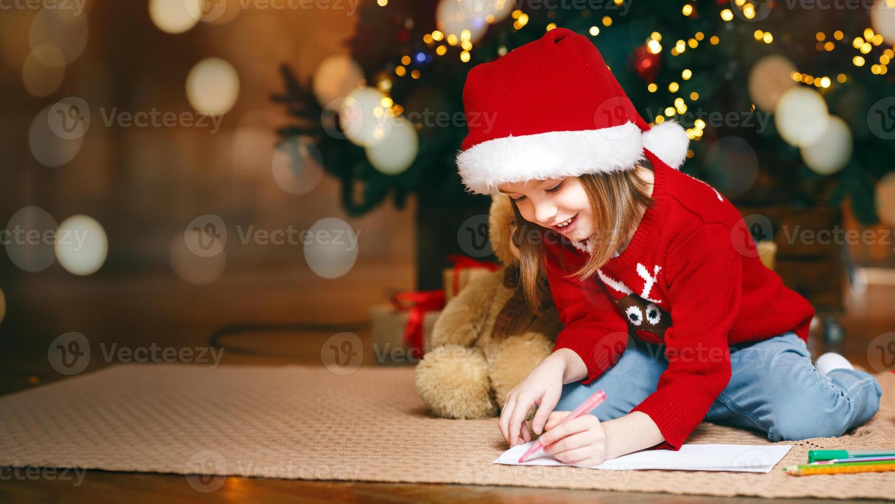 Cute little girl in red xmas hat laying on floor next to Christmas tree and writing letter to Santa, copy space photo