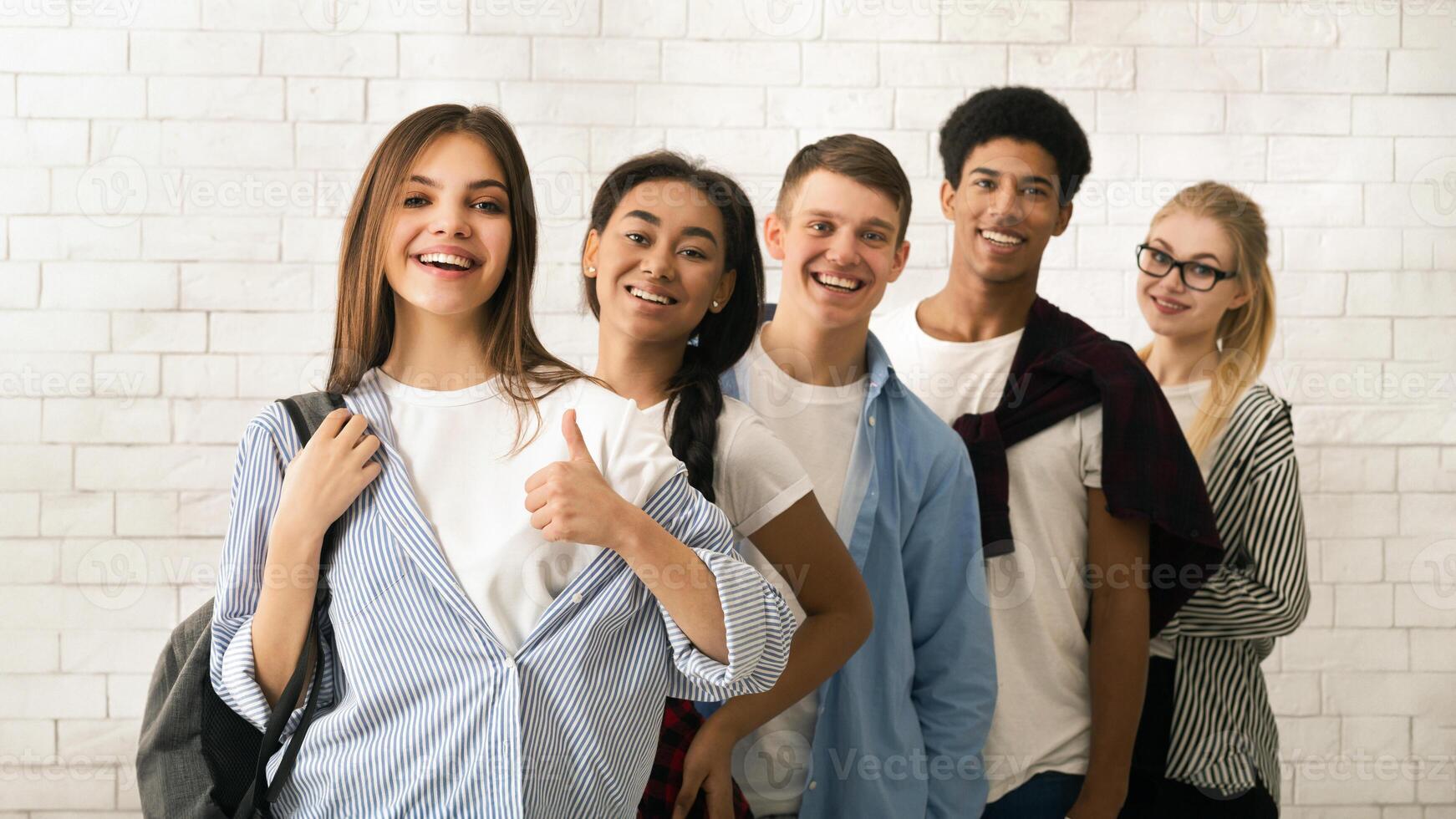 A diverse group of multiethnic teenagers is lined up, displaying a variety of cheerful expressions. The backdrop features a white brick wall, suggesting an informal or educational setting photo