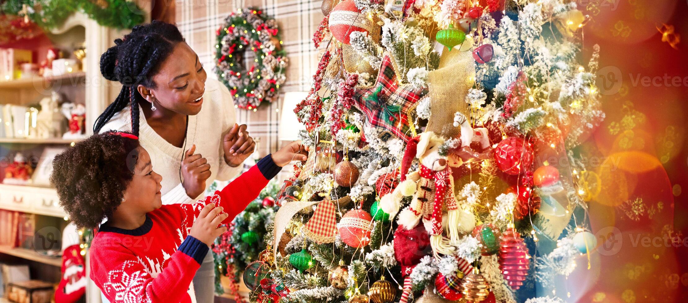 Family tradition. Cute little black girl decorating Christmas tree with her mother at home, putting on baubles and garlands photo
