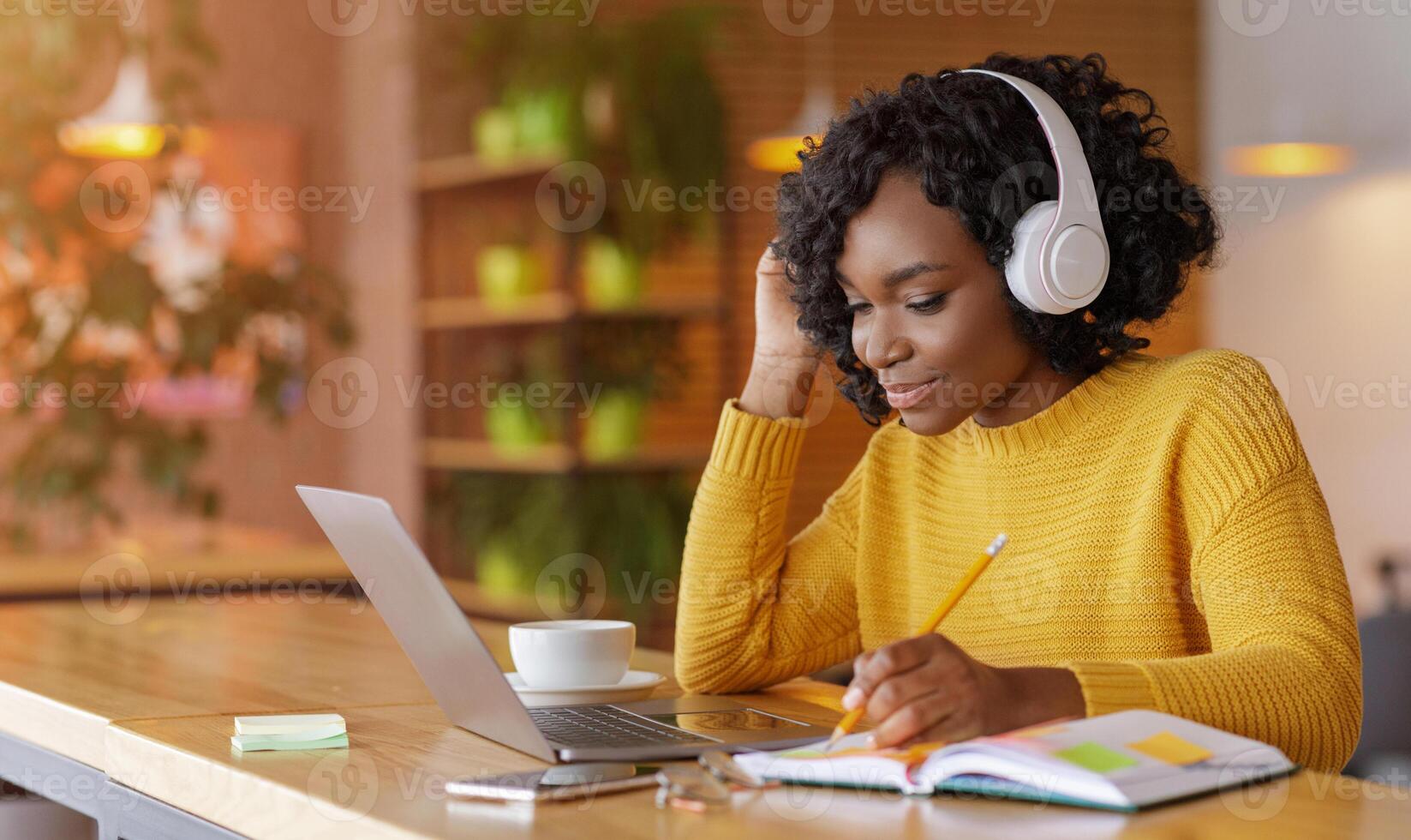 Smiling black girl with wireless headset studying online, using laptop at cafe, taking notes, copy space photo