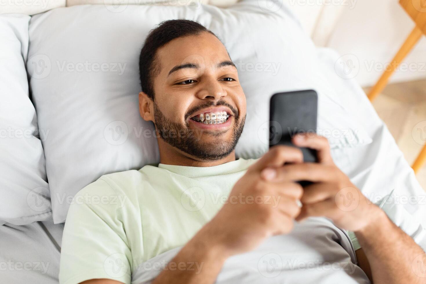 African American man is lying down in a bed, holding a cell phone in his hand. He appears to be scrolling through the screen, possibly checking messages or browsing social media. photo