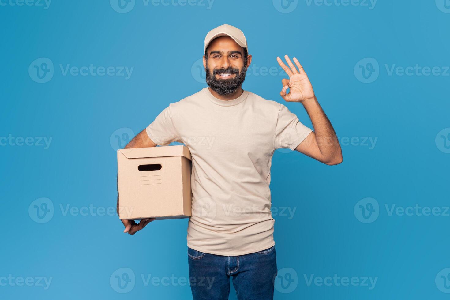 Indian delivery man dressed in a beige t-shirt and cap is standing against a blue background. He is holding a cardboard box in one hand and making an okay gesture with the other hand, smiling. photo