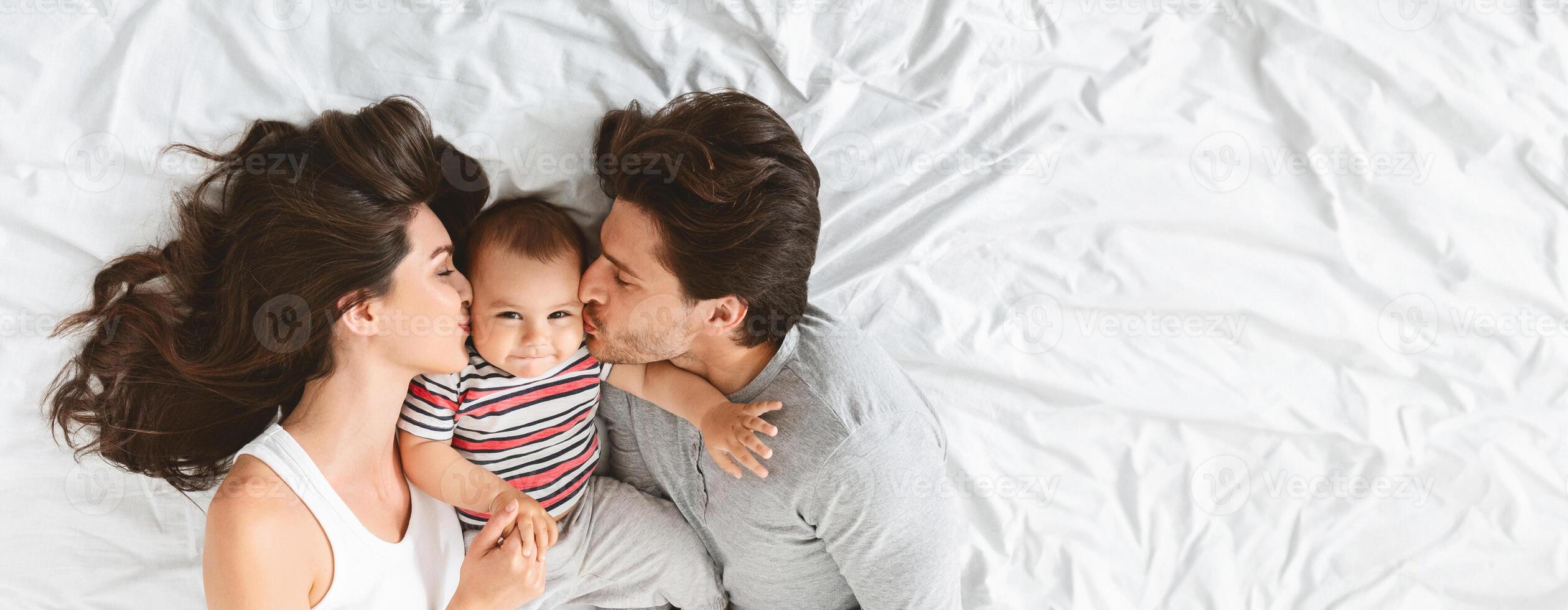 A young couple is tenderly kissing each other while holding their adorable baby in the center. They are lying on a soft white bed, radiating warmth and affection, top view, copy space photo