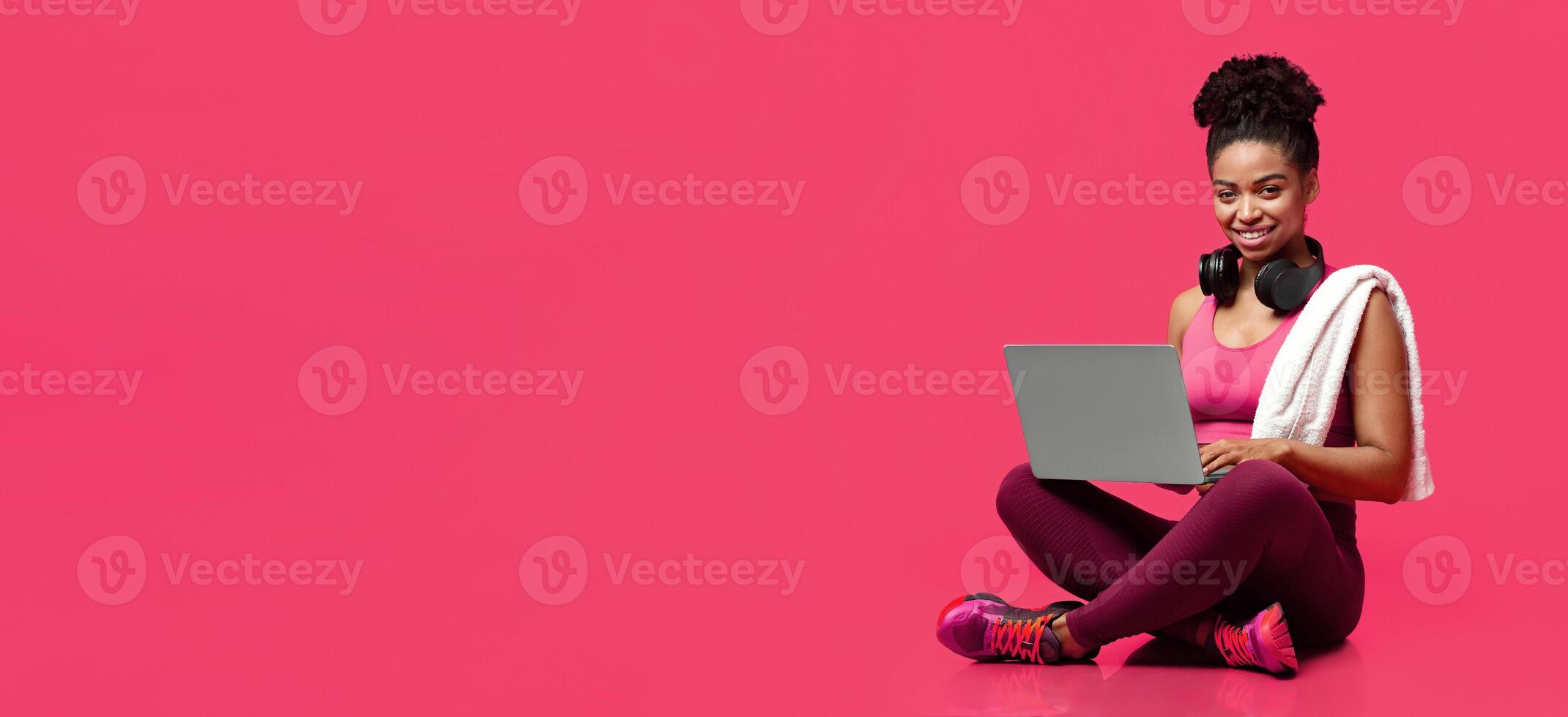 Smiling sporty athletic afro woman using laptop over red background, hanging towel and earphones, panorama with free space photo