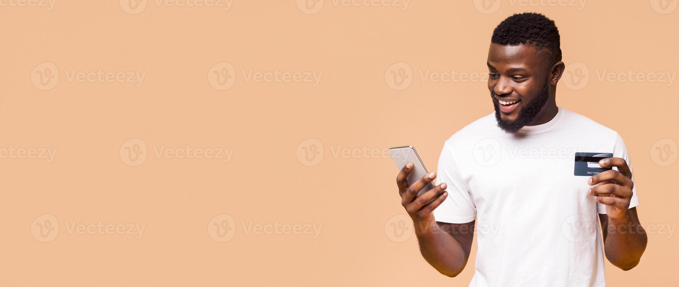 A young man with a beard smiles while holding a credit card and a smartphone, likely making an online purchase. He standing in front of a peach-colored background, copy space photo