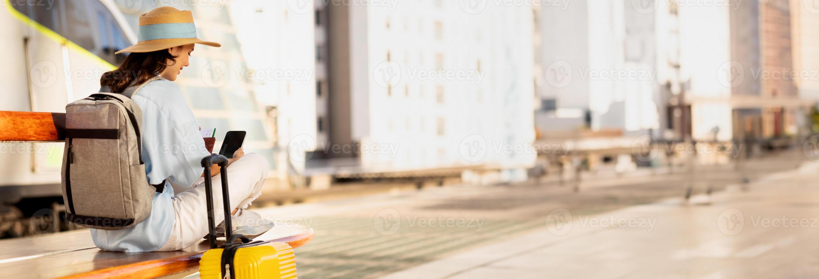 Millennial caucasian lady with backpack, suitcase, passport and ticket looks at phone with empty screen, waiting for transport on train station or airport, copy space photo