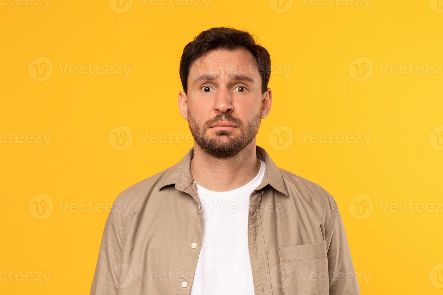 A man with a beard and brown hair is wearing a beige button-up shirt over a white t-shirt. He is looking directly at the camera with a serious expression on his face. The background is a solid yellow. photo