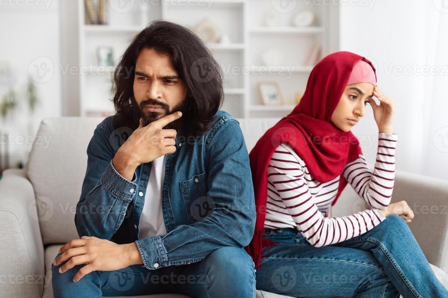 A bearded Indian man and a woman in a red hijab are sitting separately on a couch, showing expressions of concern and frustration, suggesting a difficult conversation or disagreement photo