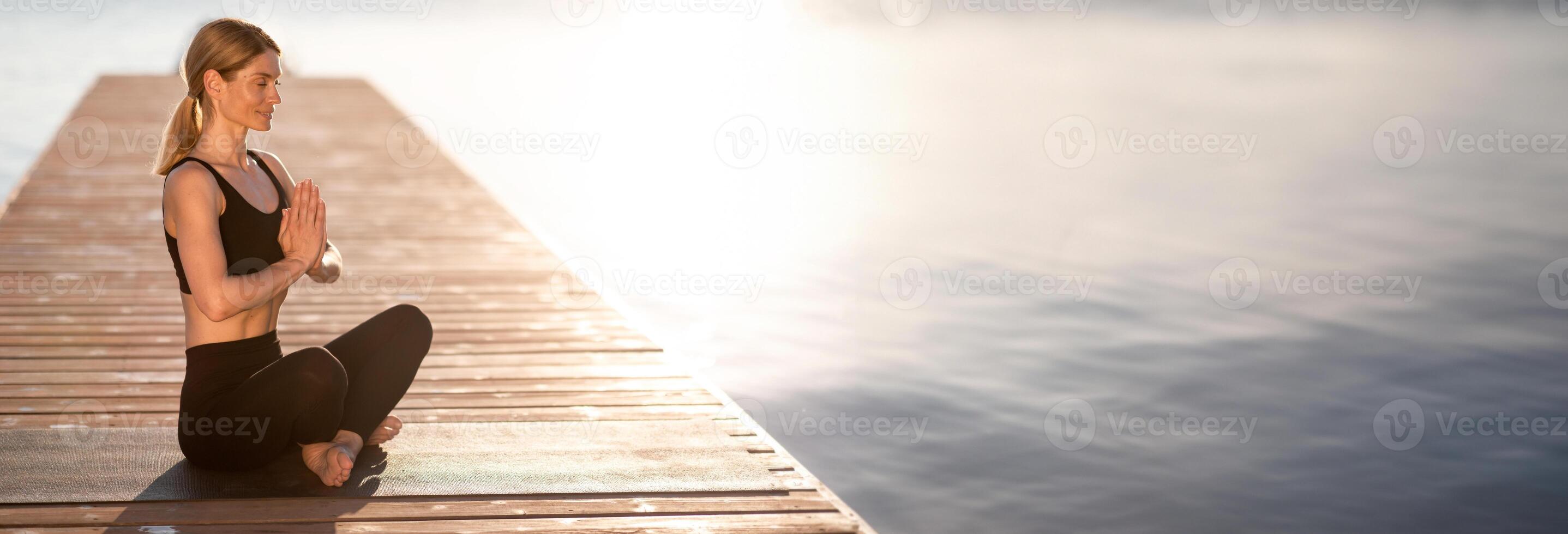 Peaceful middle aged woman meditating in lotus position on wooden pier outdoors, beautiful calm female sitting on fitness mat with closed eyes and holding hands in namaste gesture, copy space photo