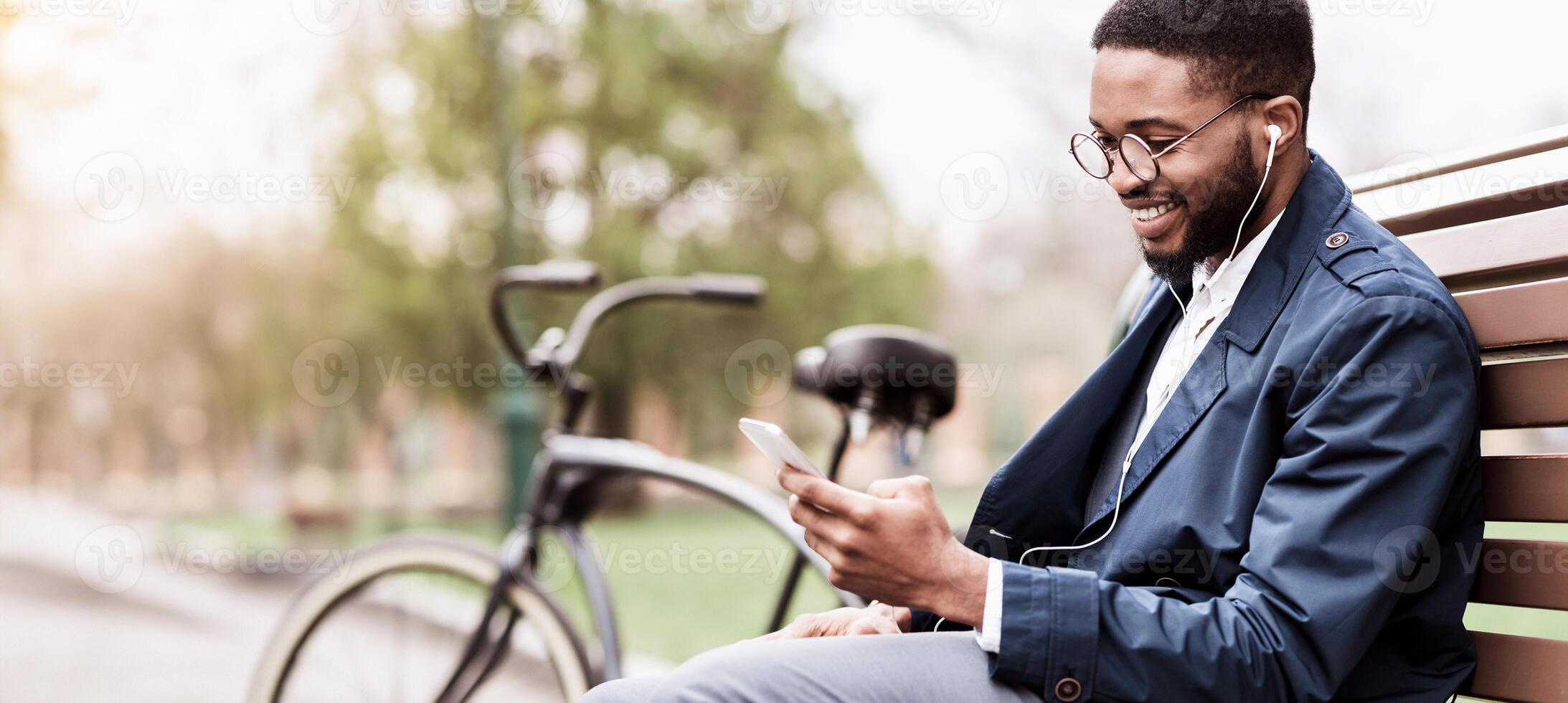 An African American man is using his phone while sitting on a bench in the park with his bicycle close by. The setting is outdoors, highlighting a peaceful yet professional atmosphere. photo