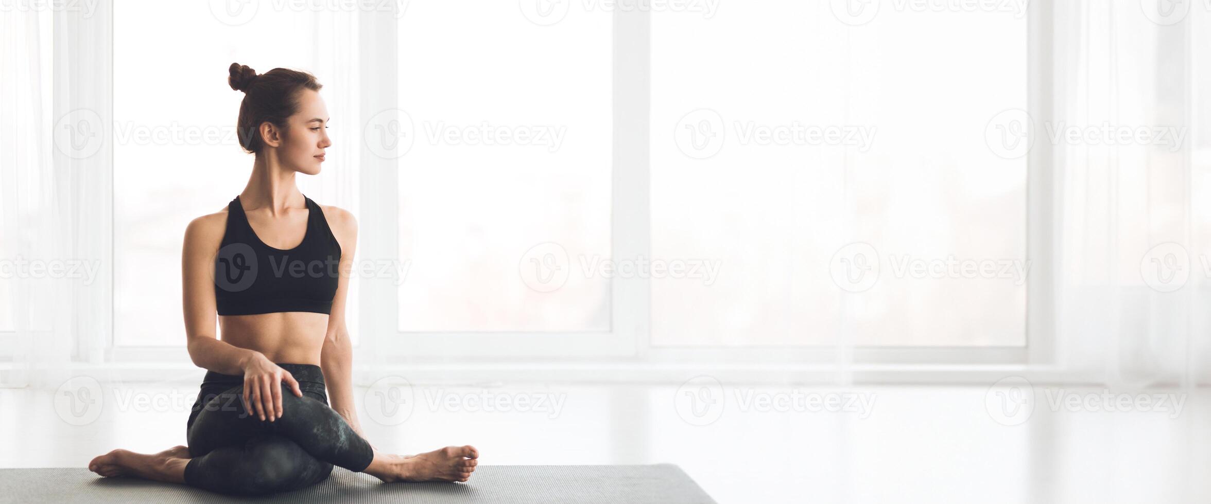 A woman is performing a seated yoga pose in a well-lit studio, focusing on her breath while surrounded by large windows, copy space photo