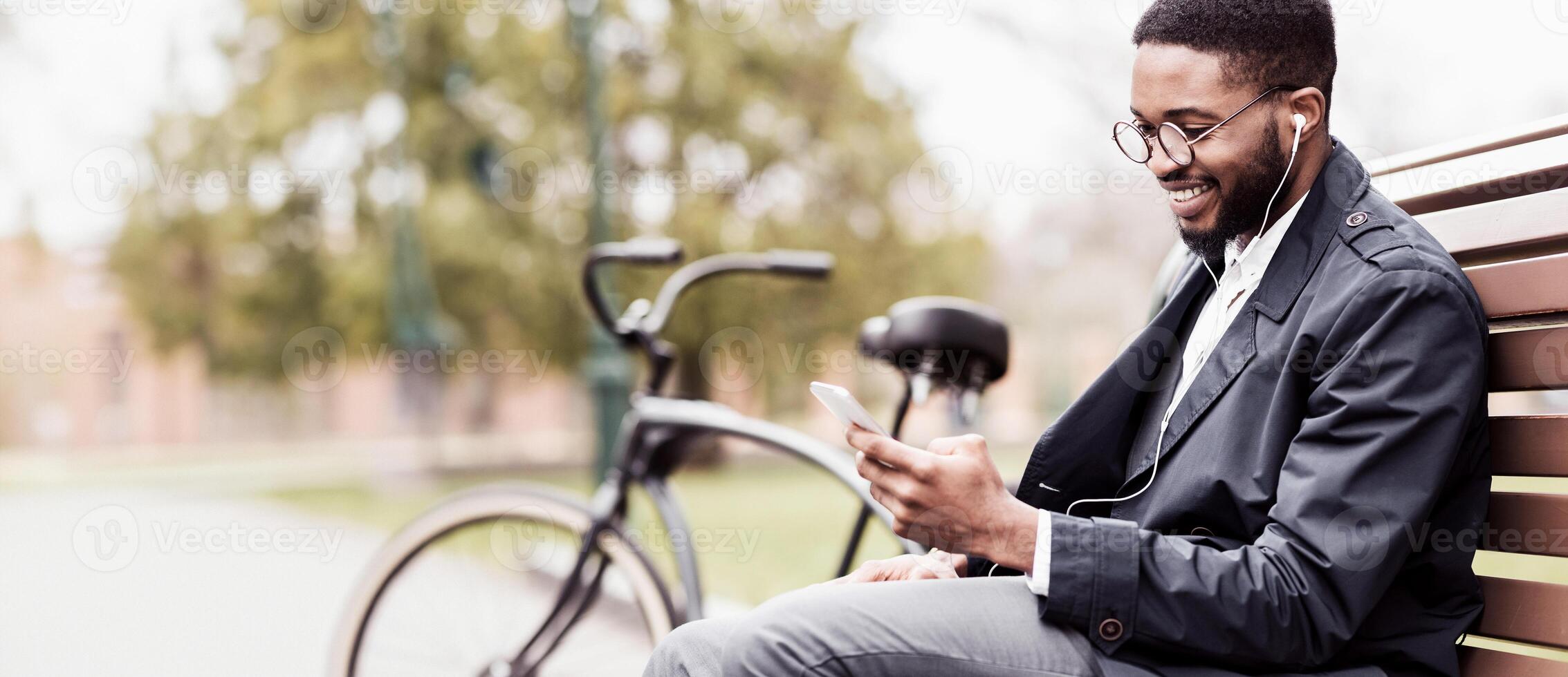A professional black man is sitting in a park with his phone and bicycle beside him. The mood is contemporary and relaxed, reflecting a modern lifestyle in a natural setting. photo