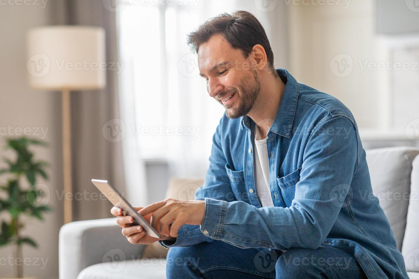 A man is sitting on a sofa in a living room, smiling and using a tablet. The sofa is a light grey color and the walls are a soft white. He is wearing a blue denim shirt and blue jeans. photo
