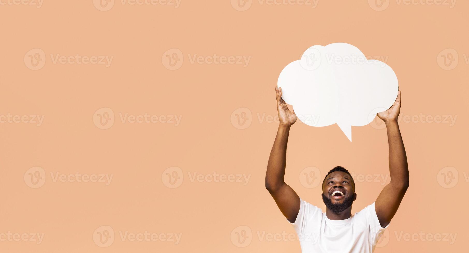 Happy Black man holding a blank speech bubble up above his head with his arms raised. He is looking up at the bubble and smiling broadly. The background is a simple peach color, copy space photo