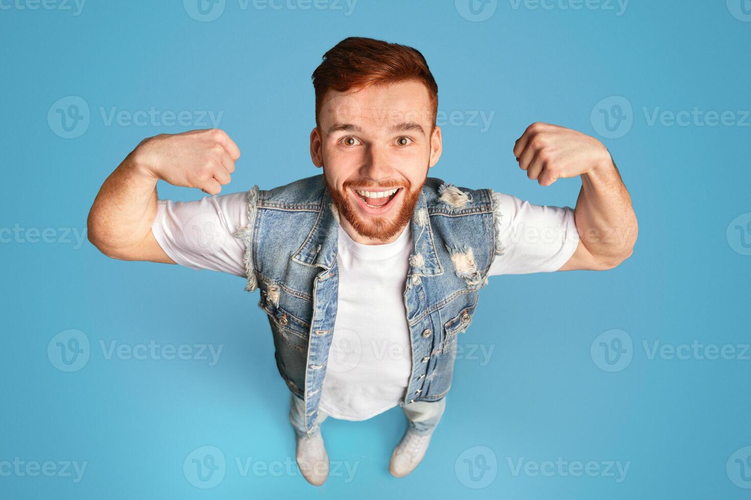 Handsome young redhead bearded man showing his power on blue background, view from above, studio shot photo