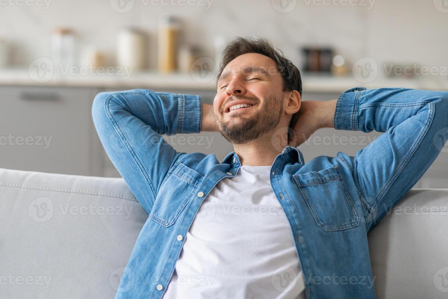 A man wearing a blue denim shirt over a white t-shirt is leaning back on a white couch with his hands clasped behind his head. He has closed eyes and is smiling. photo